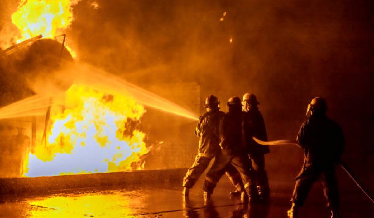 Firefighters practicing extinguishing an industrial fire at the fire station holding on to the hose spraying the fire at a storage tank.